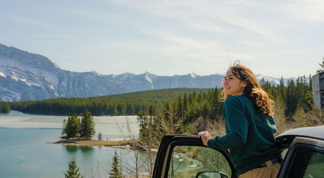 Person standing beside a vehicle overlooking a mountain lake surrounded by forest and snow-capped peaks.