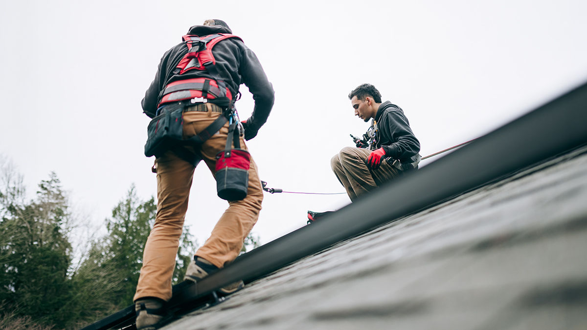 Two roofing workers wearing safety harnesses, working on a home roof during an overcast day.