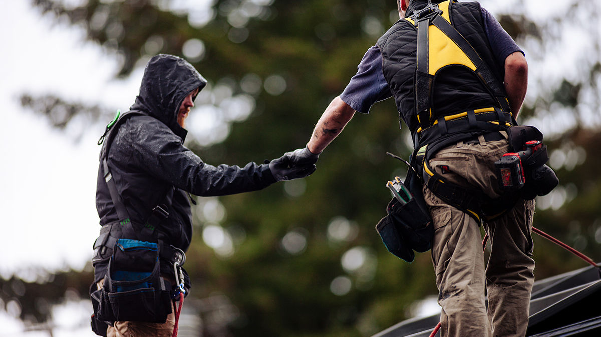 Two men in safety harnesses working on a roof in light rain, passing a tool to each other. 