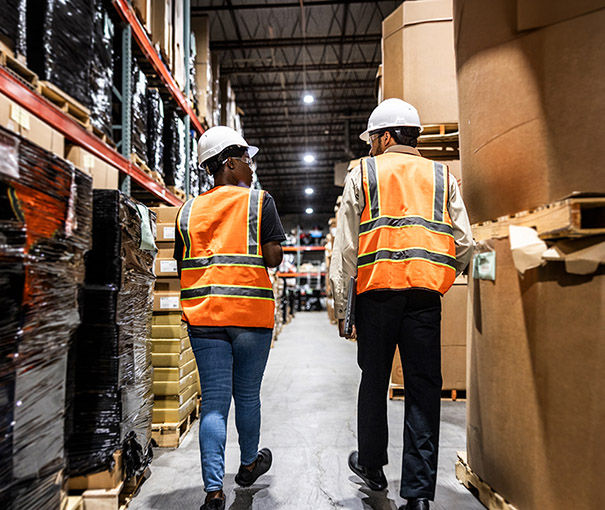 Deux ouvriers, équipés de casques et de gilets de sécurité, avancent dans une allée d’entrepôt, bordée de palettes de produits empilées.