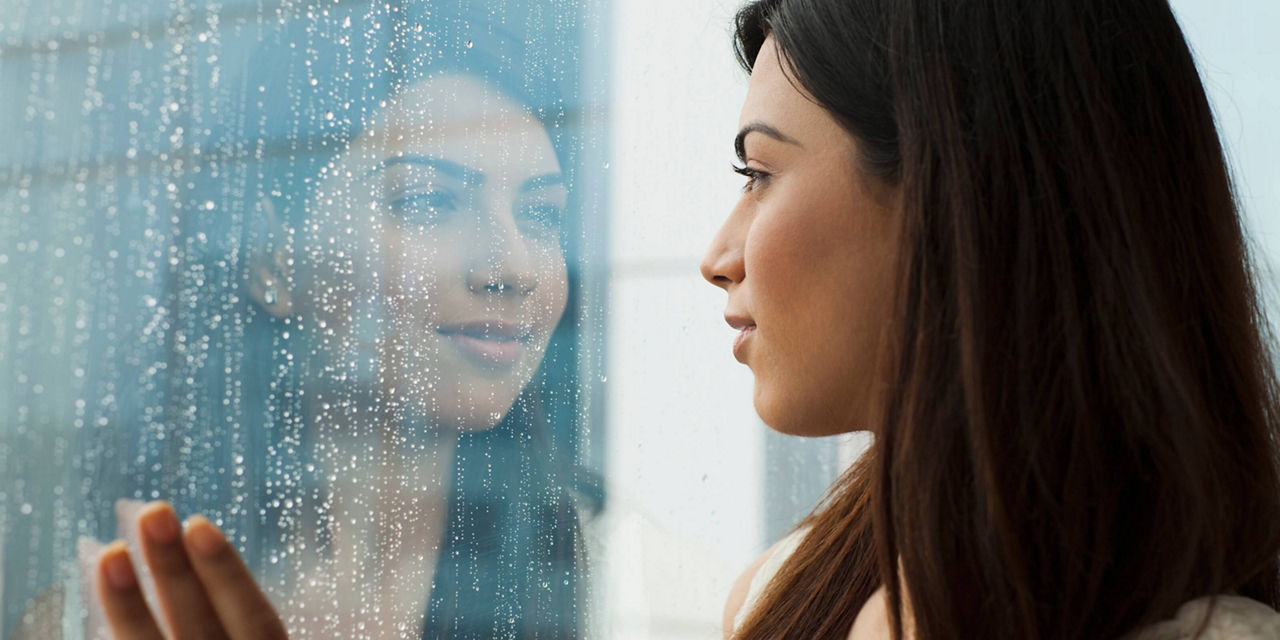 Person standing beside a rain-covered window looking outside.