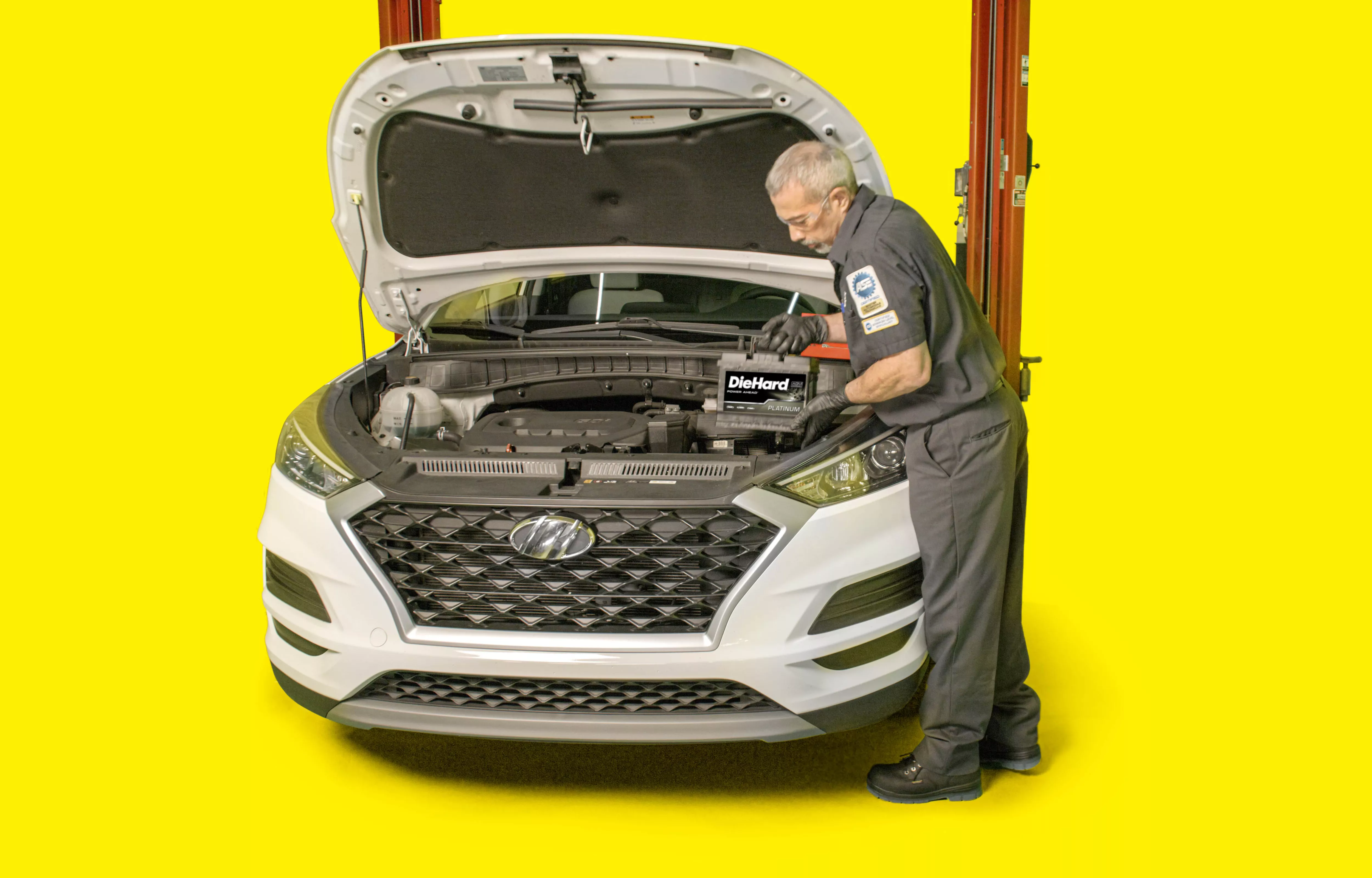 A technician installing an auto battery into a CUV with the hood popped open. The battery is labeled Die Hard.