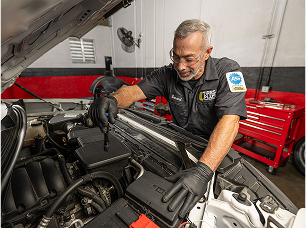 A technician in a professional garage closely inspecting a car engine bay with a flashlight.