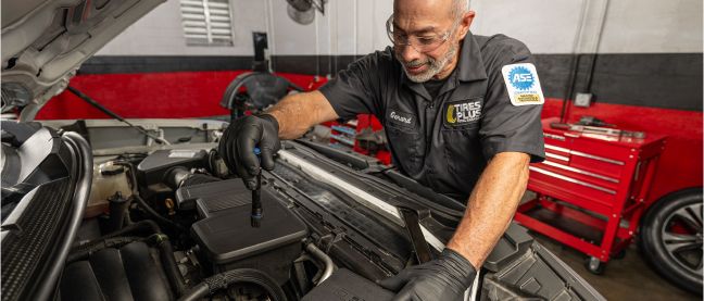 A technician in a professional garage closely inspecting a car engine bay with a flashlight.