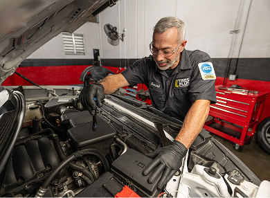 A technician in a professional garage closely inspecting a car engine bay with a flashlight.