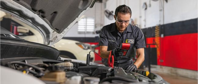 A technician in a professional garage closely inspecting a car engine bay with a flashlight.