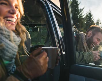smiling couple sitting in vehicle holding hot mugs with doors open in winter