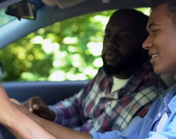 Dad looking at smiling son gripping steering wheel of vehicle