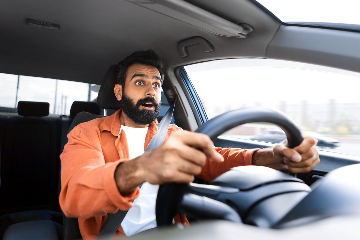 Man driving a car looking surprised whilst clenching the steering wheel with both hands