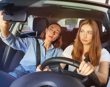 Mother illustrating how to use rear view mirror to teenage daughter in driver's seat.