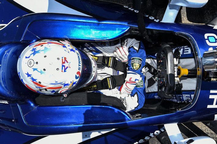 Bird's eye view of INDYCAR driver in blue race car, dressed in white and blue gear with arms crossed