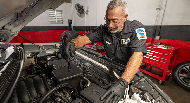 A technician in a professional garage closely inspecting a car engine bay with a flashlight.