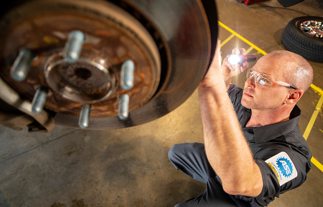 A technician closely inspecting vehicle components behind the wheel with a flashlight.