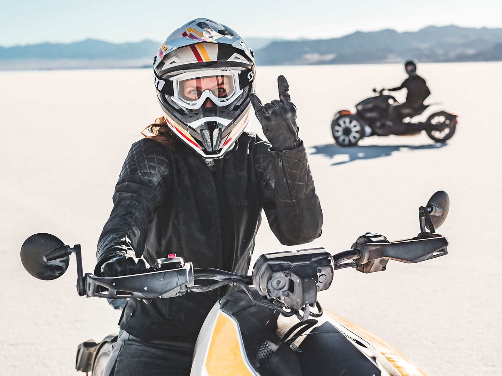 A rider gives the sign of the horns atop a Can-Am Ryker while a second rider in the background drives toward the left