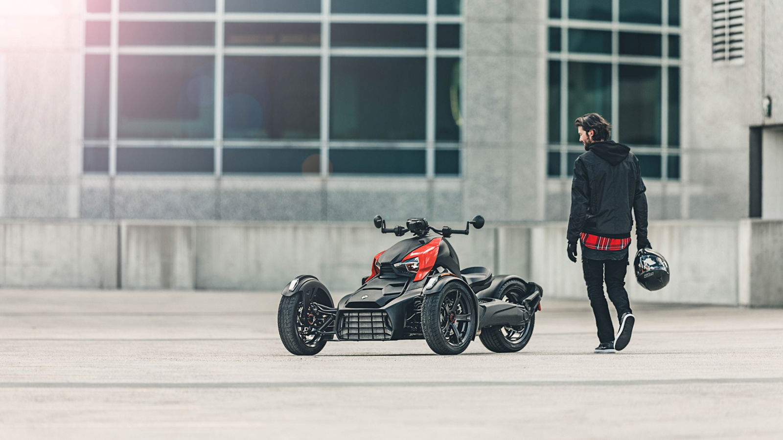 A man, helmet in hand, walking toward his Can-Am vehicle with Adrenaline Red panels parked in an empty concrete lot