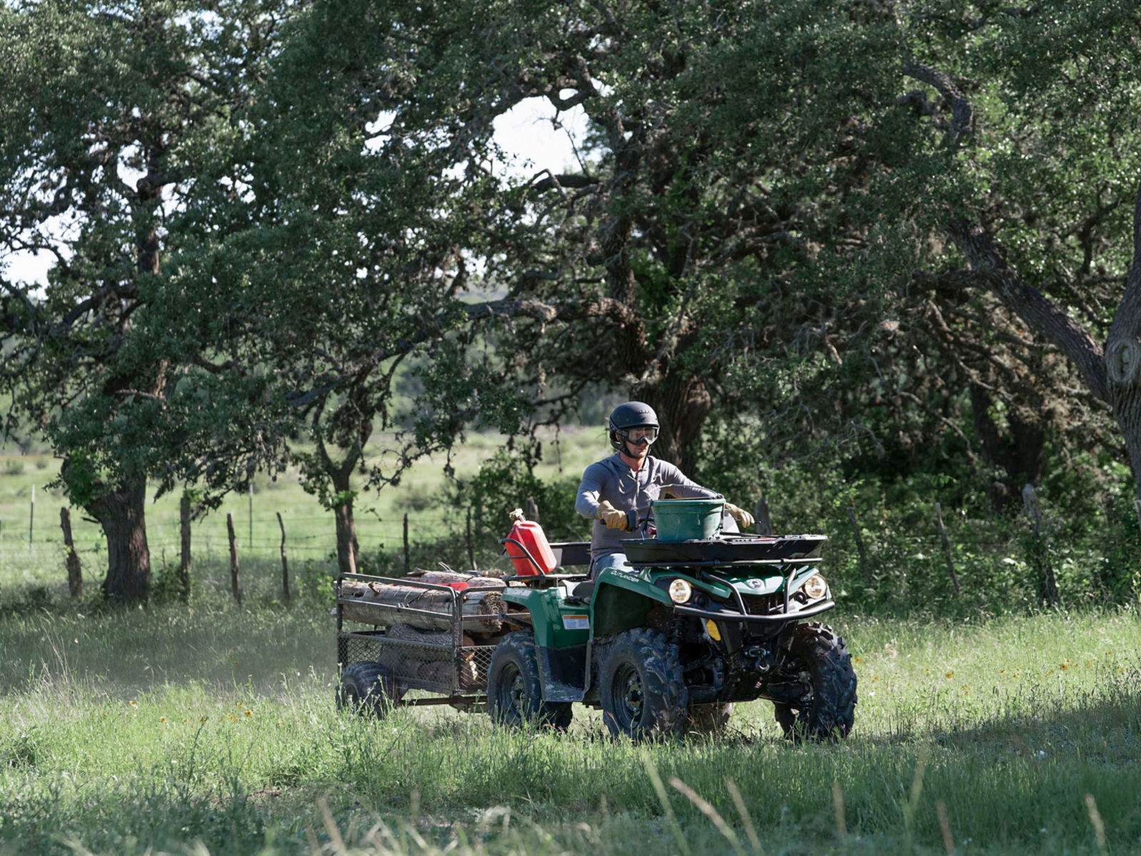 Man riding a tundra green Can-Am Outlander ATV with a trailer filled with woods