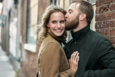 Engaged couple against a brick building - The man is kissing his bride-to-be's forehead while she looks past the camera