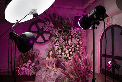 Behind the scenes photo of model sitting on couch with pink lights pointed at her and the floral background