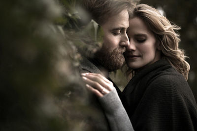 Engaged couple holding each other close - shot from behind bushes