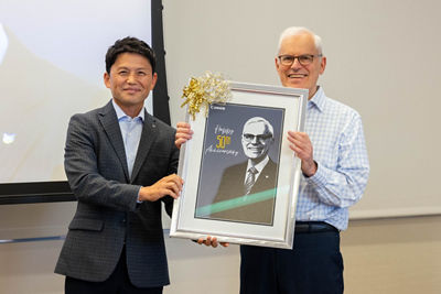 “Sammy” Kobayashi standing next to Executive Vice President Seymour Liebman who holds a portrait of him