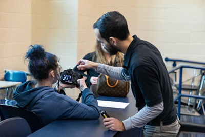 instructor pointing to camera while student looks at display panel in back