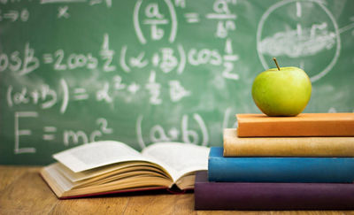 Books on a desk in front of equations on a chalkboard