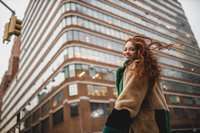 Curly red-haired model smiling over her shoulder in front of a large glass building