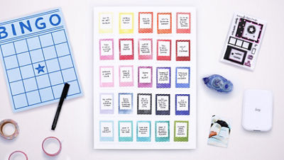 Flat lay of a blank bingo board, colorful aspiration cards stamped with multicolor striped frames, and craft supplies on a white background including washi tape, pens, the Canon Ivy Mini Photo Printer, adhesive runner, and ‘Captured’ Stamp Set by The Paper Curator