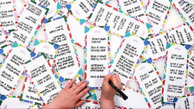 Person writing "read 15 books" in black marker on colorful bordered notepad sheets spread across a table, creating a list of aspirations for this project such as “print more photos,” “try a new coffee shop,” and “have a dance party in pajamas.