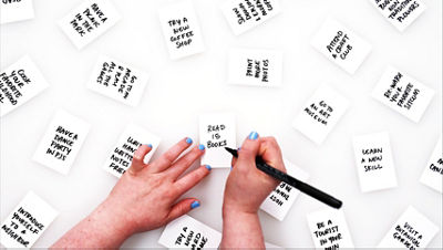 Hands writing “Read 15 books” on a small white card surrounded by other aspiration cards like “Try a new coffee shop,” “Attend a craft club,” and “Go to an art museum.”