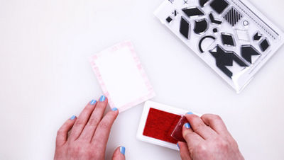 Hand with blue nail polish pressing a clear stamp into a pink ink pad next to a small cardstock card with a pink stamped border and the Pattern Play Stamp Set by The Paper Curator.