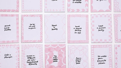 Overhead view of multiple small cardstock aspiration cards arranged in rows. Each card has a pink stamped decorative border and a handwritten aspiration in the center, such as “go to an art museum,” “read 15 books,” and “host a dinner party.”