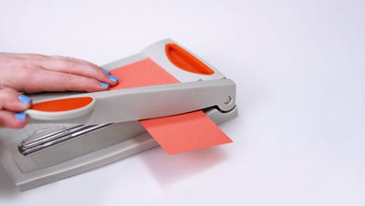 Close-up of a hand with blue nail polish using a paper trimmer to cut a strip of orange cardstock paper into a smaller rectangular piece sized 2 1/2 by 3 1/2 inches.