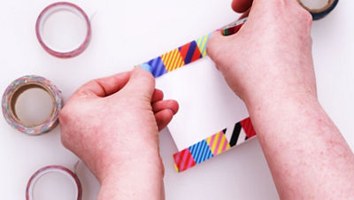 Close-up of hands placing a strip of bright, patterned washi tape along the edge of a small rectangular cardstock card to create a decorative border.