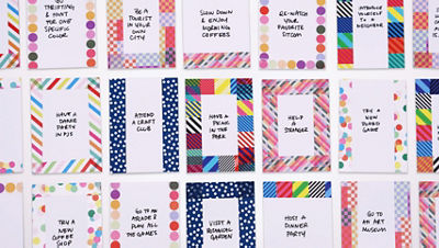 Overhead view of multiple small cardstock aspiration cards arranged in rows. Each card has colorful decorative washi tape borders and a handwritten aspiration in the center, such as “attend a craft club,” “have a picnic in the park,” and “help a stranger.”