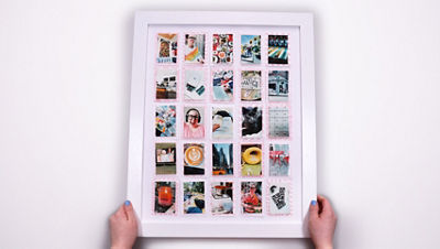 Hands hold a white frame containing a completed photo bingo board, where each square of the grid has been replaced with a small printed photo documenting completed aspirations.