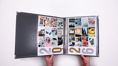 Hands hold an open binder-style scrapbook album with two pages of small printed photos arranged in grid layouts, offering an alternative way to store and display photos from this completed project.