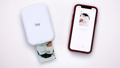 The Canon Ivy 2 Mini photo printer prints a small photo of an open book while a nearby smartphone screen shows the same image processing in the Canon Mini Print app.