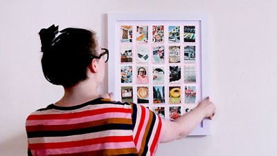 A person with glasses and a striped shirt hangs a white frame on a white wall containing a completed bingo board grid of small photos with pink stamped borders.