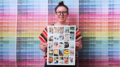 A person stands in front of a colorful color swatch wall holding a finished photo bingo board displaying a grid of small printed photos.