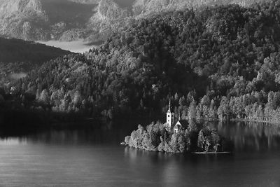 Black and white landscape image of a steepled building on a small island in the middle of a lake with mountains in the background