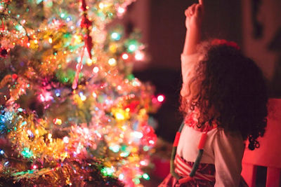 young girl sitting in a chair next to a lit Holiday Tree pointing up