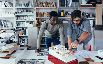Two men in an office lean over a desk and look at a small building model