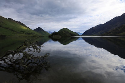 Wide-angle shot of a lake with green mountains in the backgroud and clouds reflecting in the water