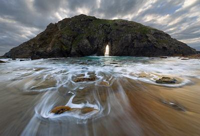 Image of Arch rock in Oregon framed by strong grey clouds above and shallow water below flowing over sand and occasional smooth stones