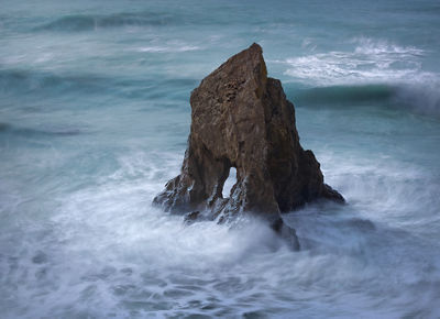 One large dark rock jutting up out of the foamy ocean waves with a piece missing from the center