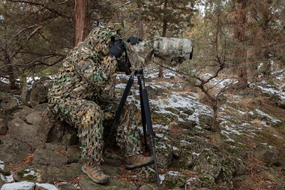 Image of photographer looking through a camera on a tripod with a ghillie camouflage suit