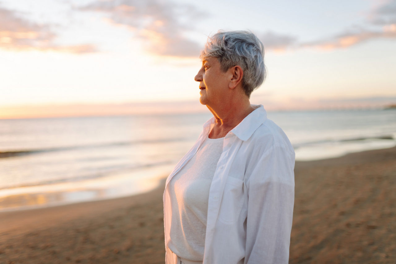 An elderly woman strolls barefoot along a sandy beach at sunset. A gray-haired woman enjoys the sunrise by the seashore, feeling free. Concepts of retirement, freedom, and relaxation.
