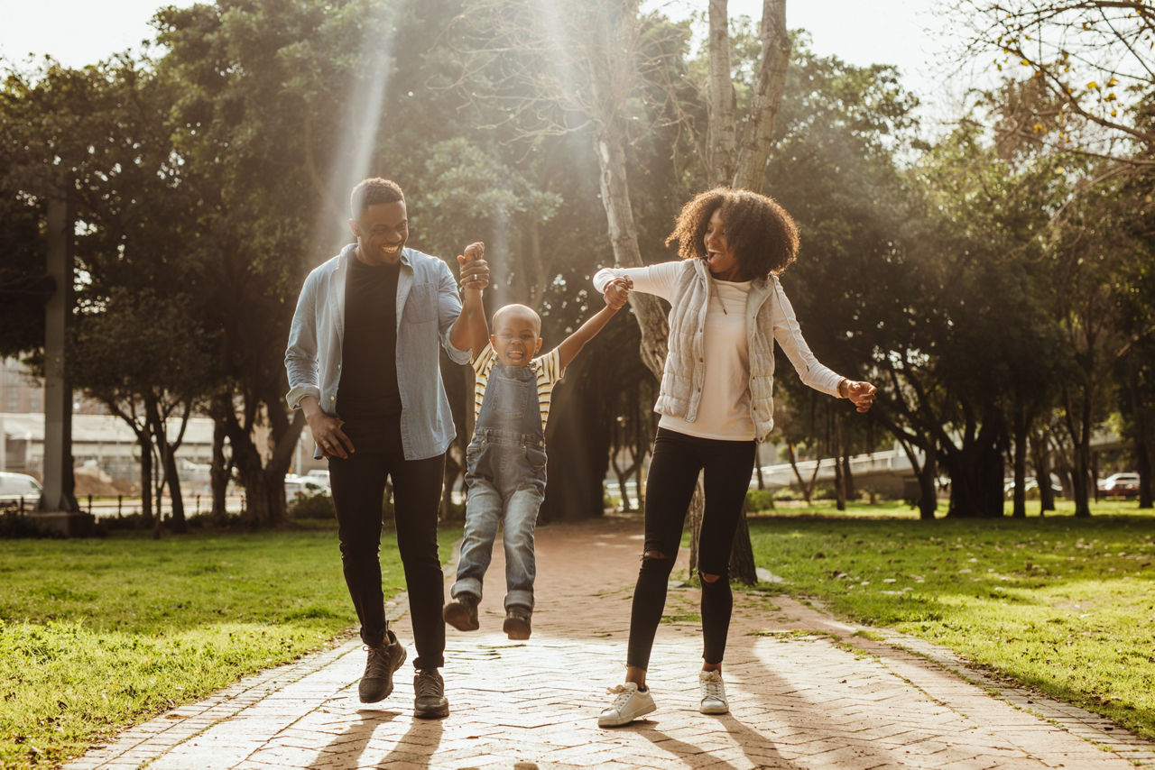 A Black couple holding their child's hands, swinging them playfully, walk down a sun-dappled path in a park. Their joyful expressions highlight a happy family outing.