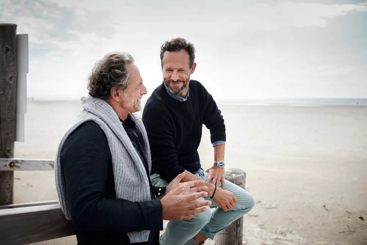 Two men, one older, one younger, are engaged in a friendly conversation on a beach. Both are smiling, suggesting a warm interaction amidst the casual, outdoor setting.